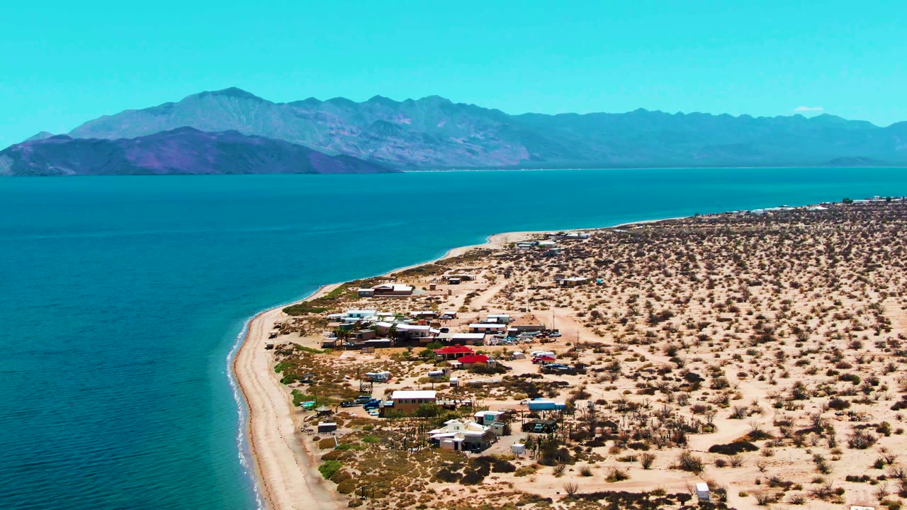 At 0:02, the aerial view shows Bahia De Los Angeles water and beach running diagonally through the frame, with scattered shore buildings, sandy desert lots, mountain ridges, and no visible people.