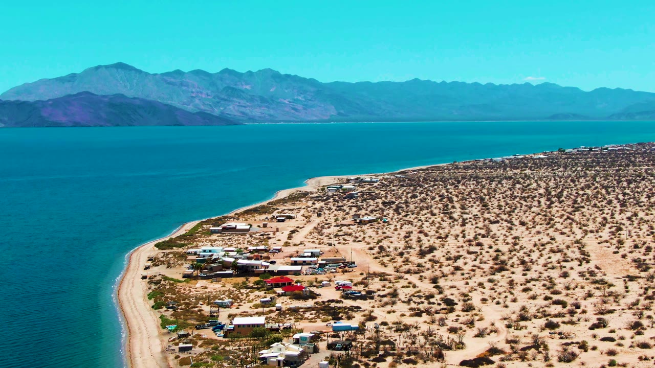 Aerial view over Bahia De Los Angeles with blue water at left, a sandy shoreline, desert camp structures and vehicles below, distant mountains, and no visible people.
