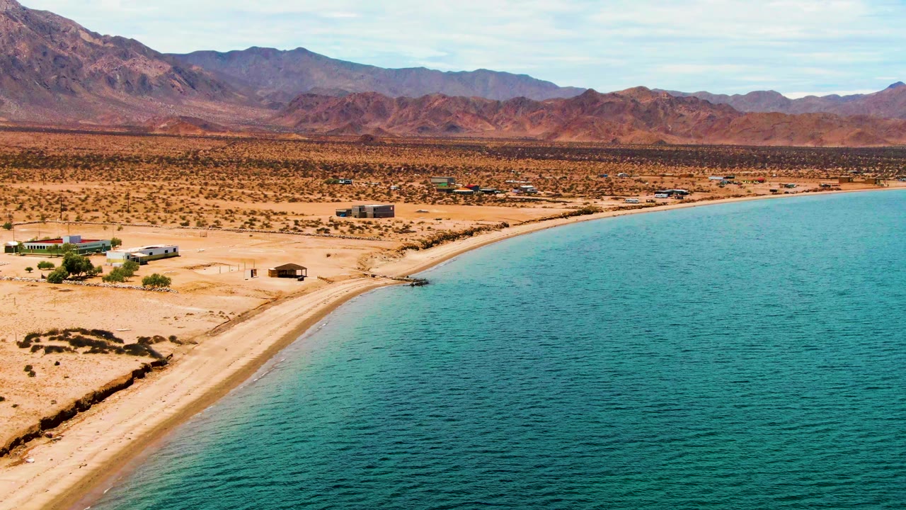 Aerial view in Bahia De Los Angeles, Baja California Sur, Mexico, showing a curved sandy shoreline, turquoise water, sparse desert buildings, distant mountains, and a tiny person near the beach.