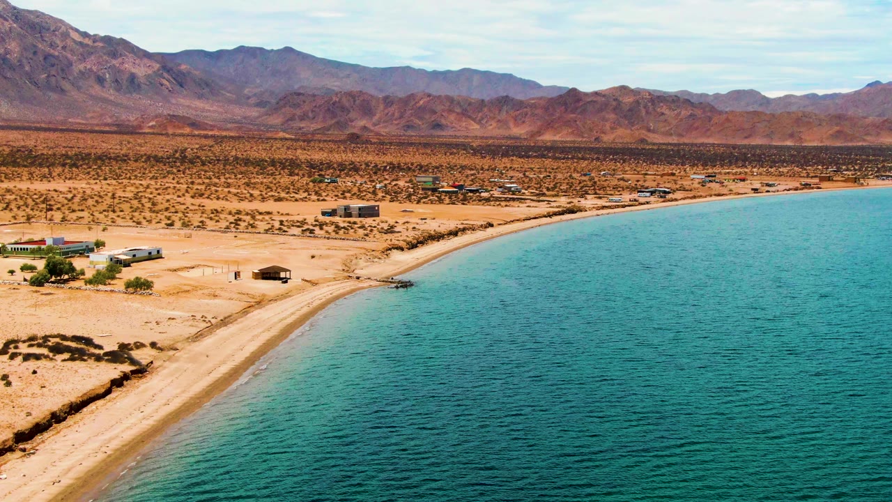 Wide aerial frame in Bahia De Los Angeles with turquoise water on the right, a sandy beach curving through the center, small inland structures, distant mountains, and a tiny person along the shore.