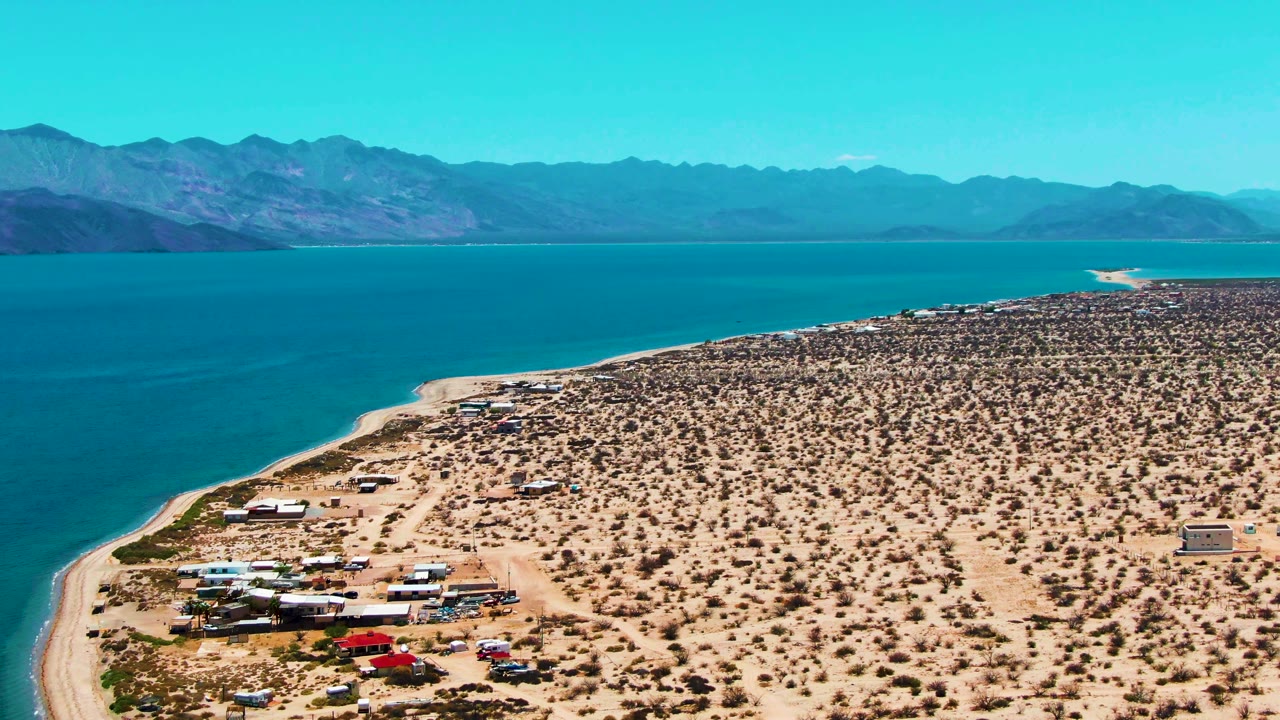 Aerial view over Bahia De Los Angeles with blue water on the left, sandy desert and sparse shrubs on the right, vehicles near the shore, distant mountains, and no visible people.