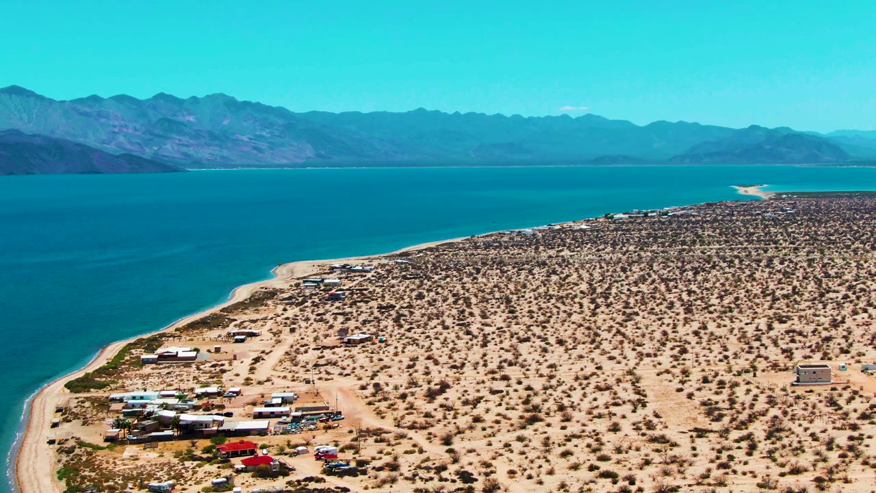 High aerial view of a curved desert coastline with RVs, cars, blue water, and mountains.
