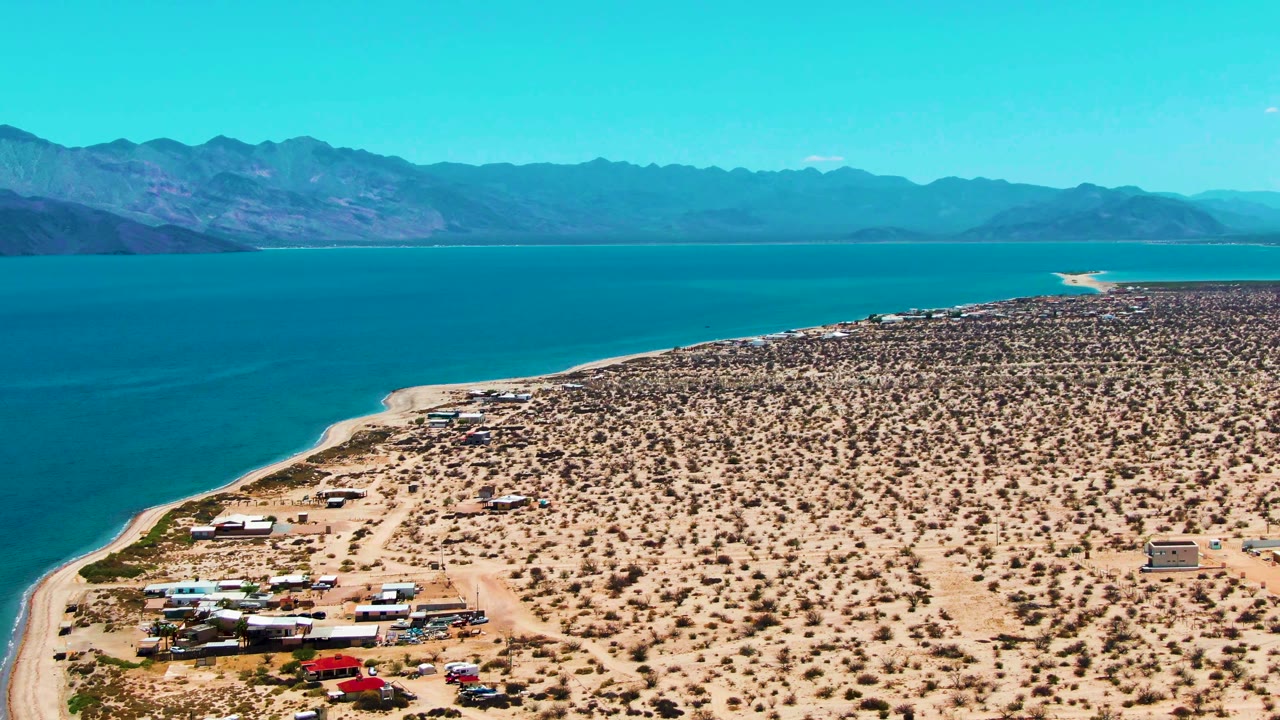 Aerial view of Bahia De Los Angeles with turquoise-blue water along the left edge, a sandy road and vehicles near the shoreline, broad desert scrubland, distant mountain ridges, and no visible people.