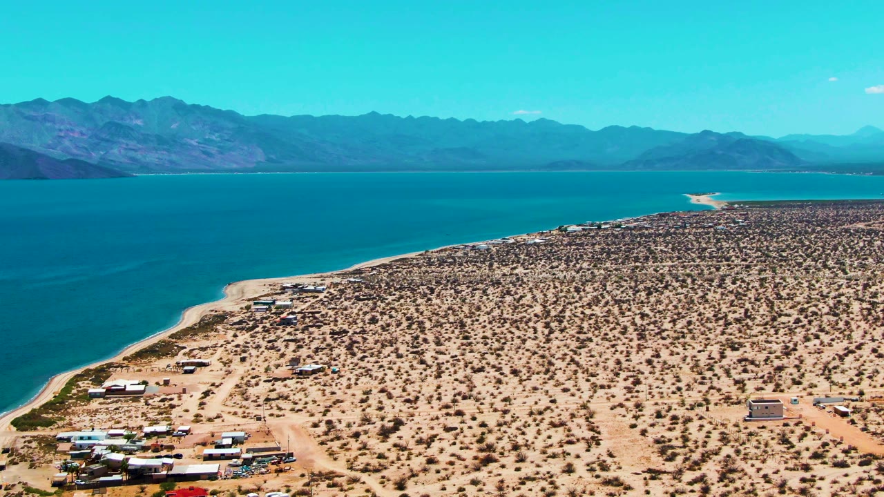 Opening aerial frame of Bahia De Los Angeles with blue water along a sandy desert shoreline, sparse scrub, distant mountains, RVs and small beach structures, and no visible people.