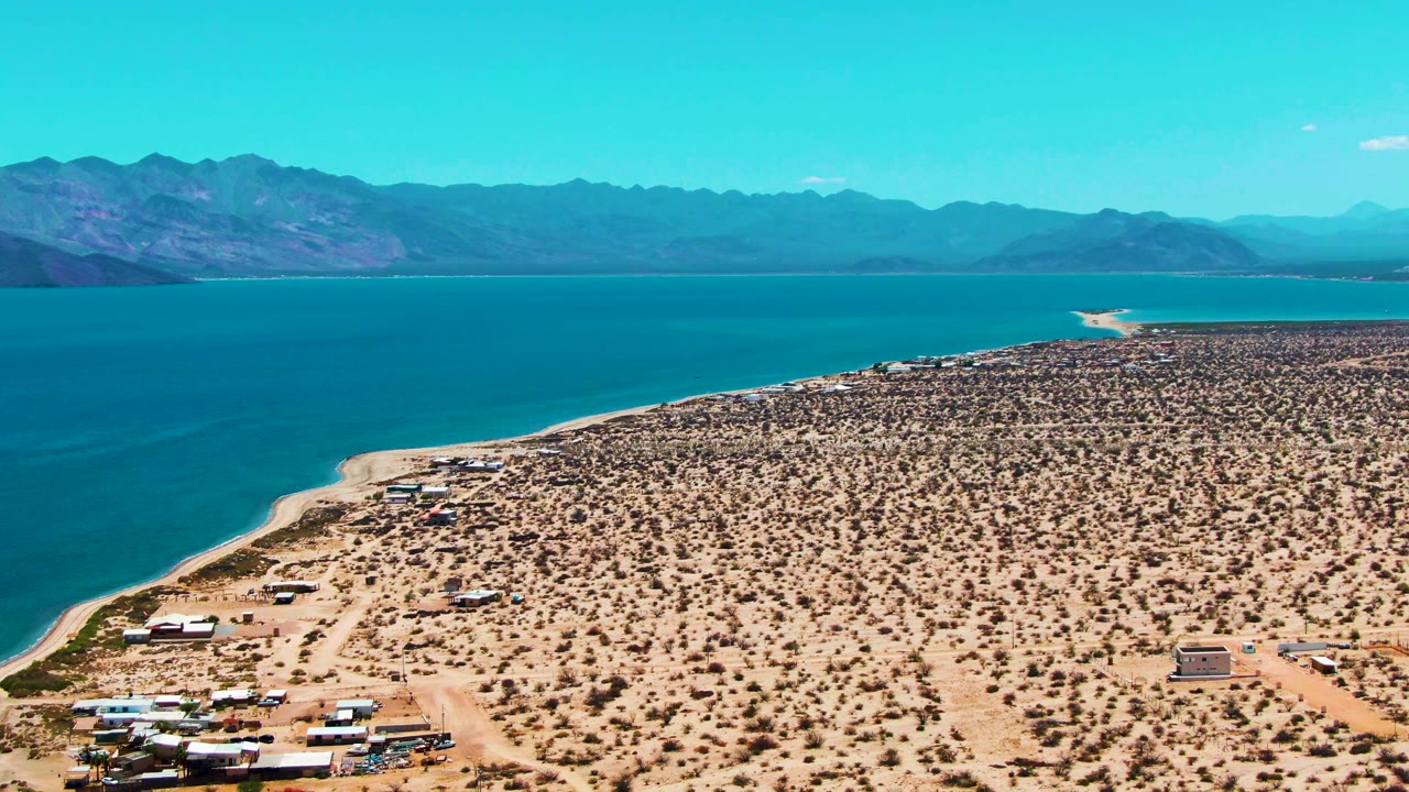 Wide aerial shoreline with turquoise water, arid scrubland, beach RVs, and mountain horizon.
