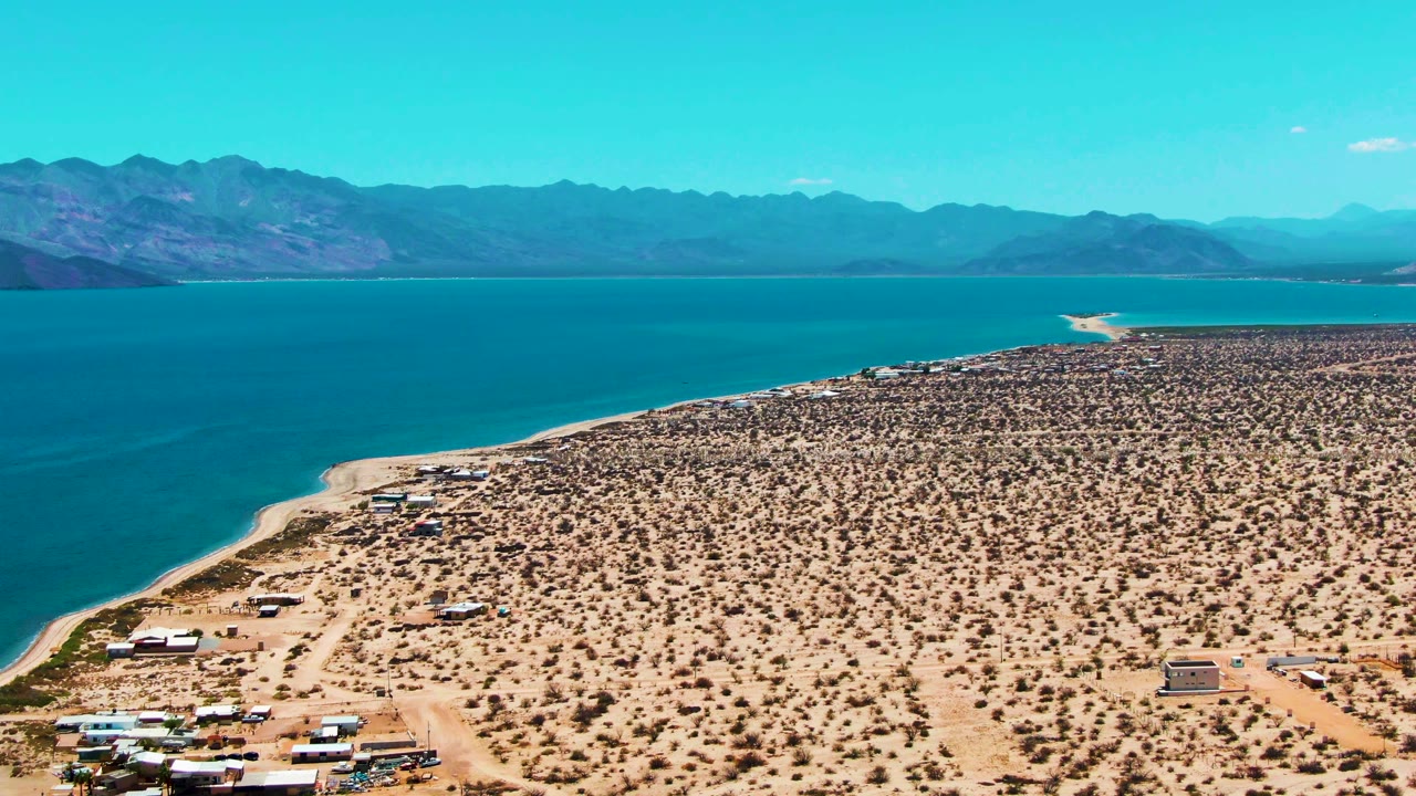 Third frame of Bahia De Los Angeles with blue bay water filling the left side, a sandy beach edge, dry scrubland, scattered structures, distant mountains, and no visible people.
