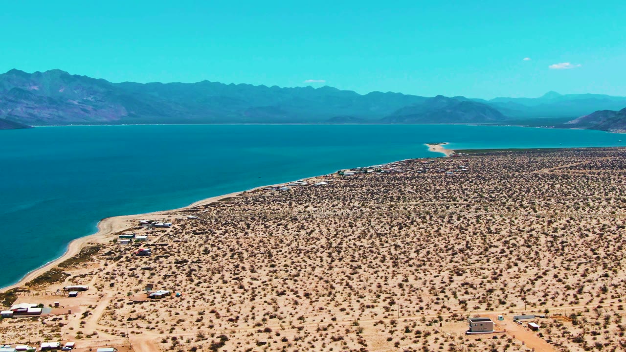 Wide aerial view over Bahia De Los Angeles desert shoreline, with sparse scrub, dirt tracks, blue water, distant mountains, and no visible people.