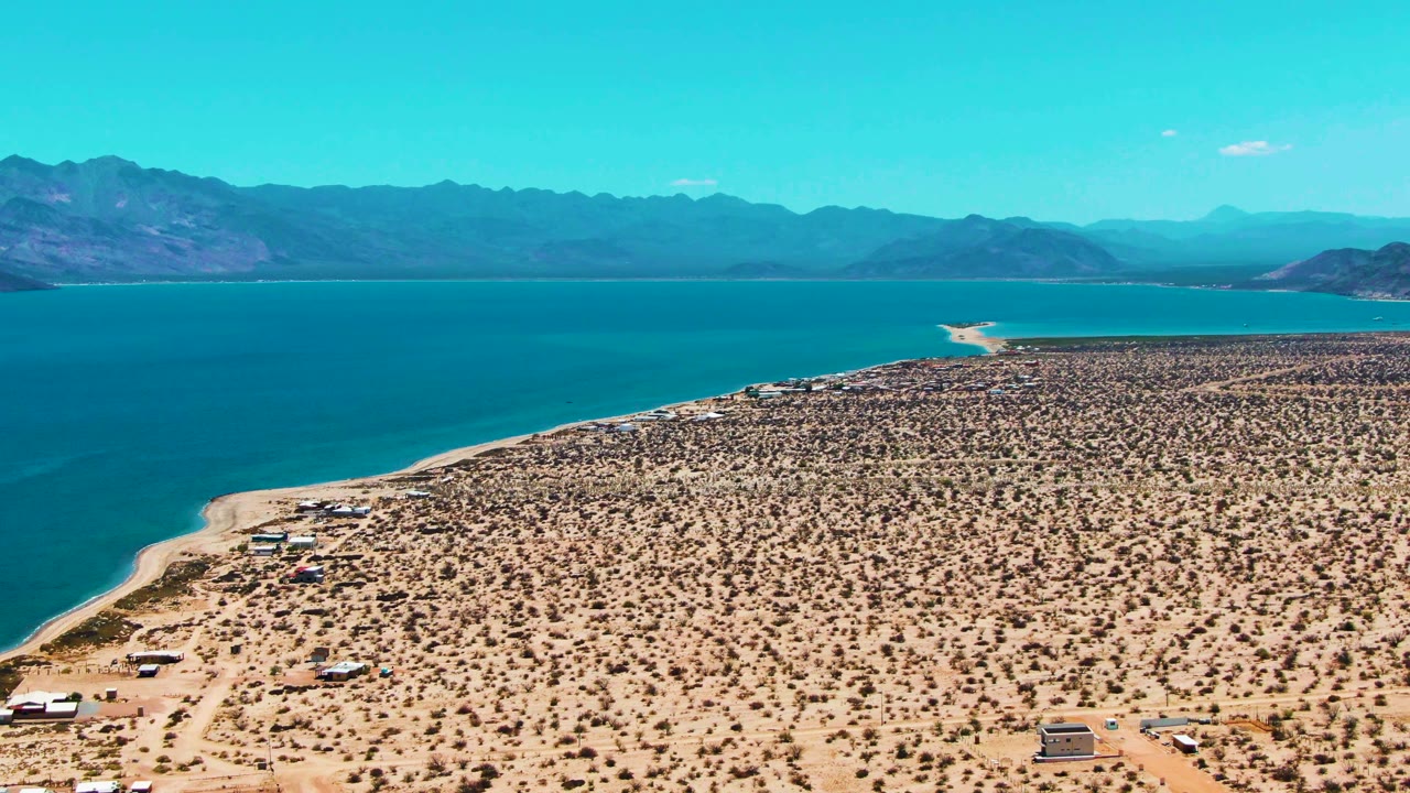 Aerial frame of Bahia De Los Angeles showing tan desert terrain, a curving shore, open blue water, mountain ridges, and no visible people.