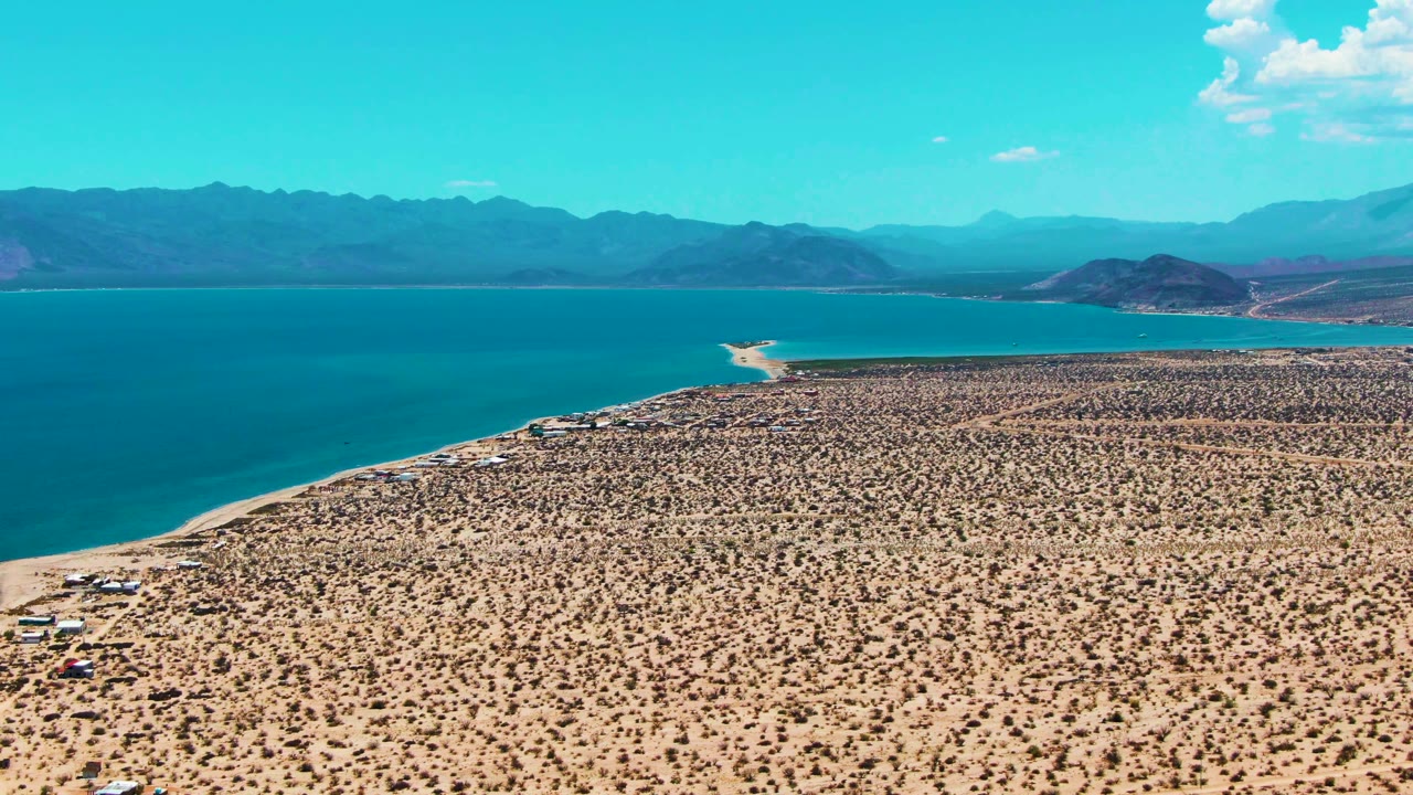 Opening aerial frame over Bahia De Los Angeles showing tan desert scrub, blue water on the left, distant mountains, scattered clouds, small shoreline structures, and no visible people.