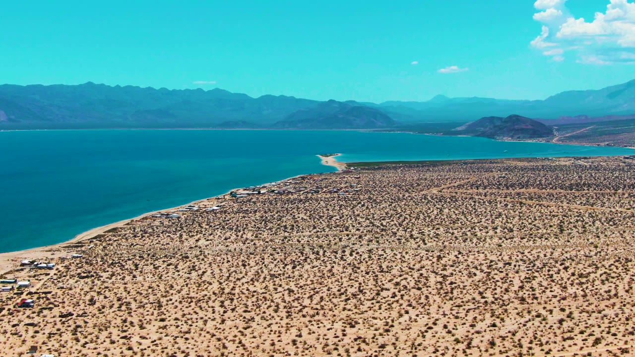 Aerial frame over Bahia De Los Angeles with a broad tan desert foreground, the bay curving along the left edge, hazy mountain ridges in back, and no visible people.