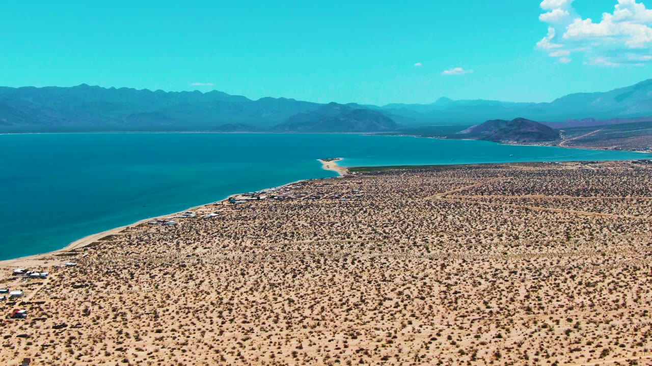 Mid-clip aerial view of Bahia De Los Angeles showing dry scrubland across the foreground, calm blue water to the left, a pale sand spit near the middle distance, mountains, and no visible people.