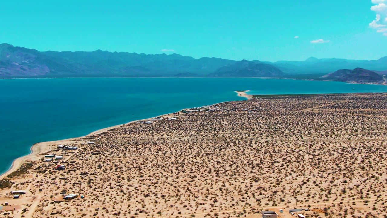 A wide aerial frame over Bahia De Los Angeles shows sandy desert scrub, a curving blue shoreline, scattered small structures near the beach, distant mountains, and no visible people.