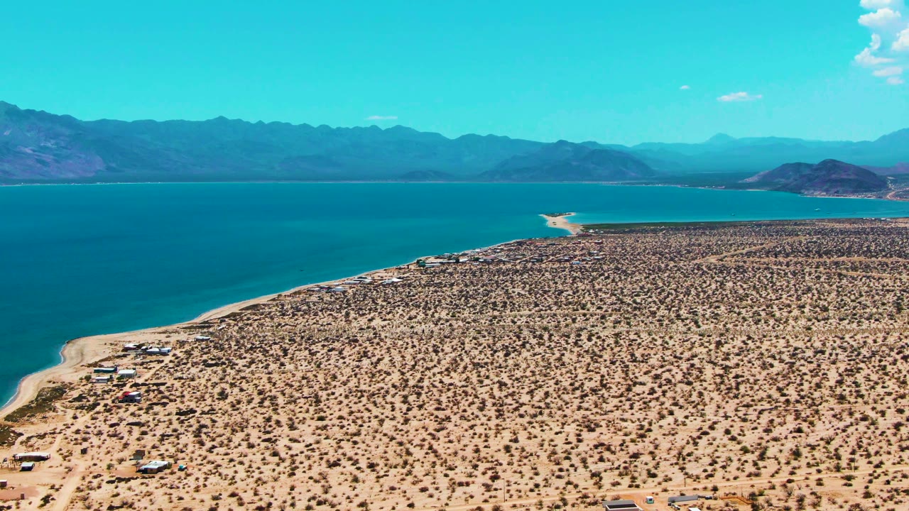 At 0:01, the aerial view holds over Bahia De Los Angeles with the desert foreground slightly shifted, small tents or vehicles near the beach, blue water, distant mountains, and no visible people.