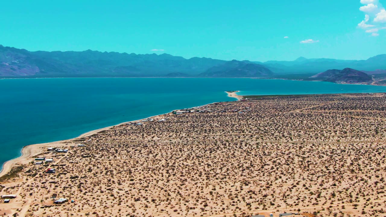 At 0:02, Bahia De Los Angeles appears from above with a sandy scrub plain, a pale beach edge, small shoreline structures, saturated blue water, layered mountains, and no visible people.