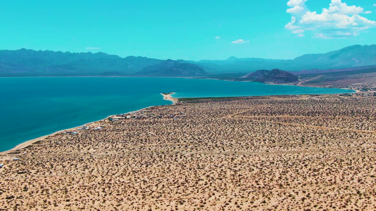 Wide aerial frame from Bahia De Los Angeles with tan desert brush below, turquoise water along the shore, layered mountains in haze, and no visible people.