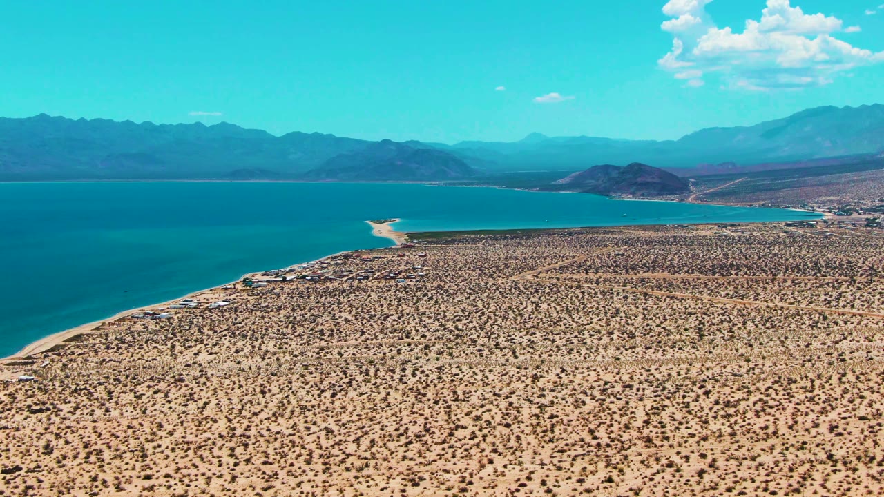 Aerial view of Bahia De Los Angeles with a broad desert plain, blue bay water, pale sandy edge, distant mountains, and no visible people.