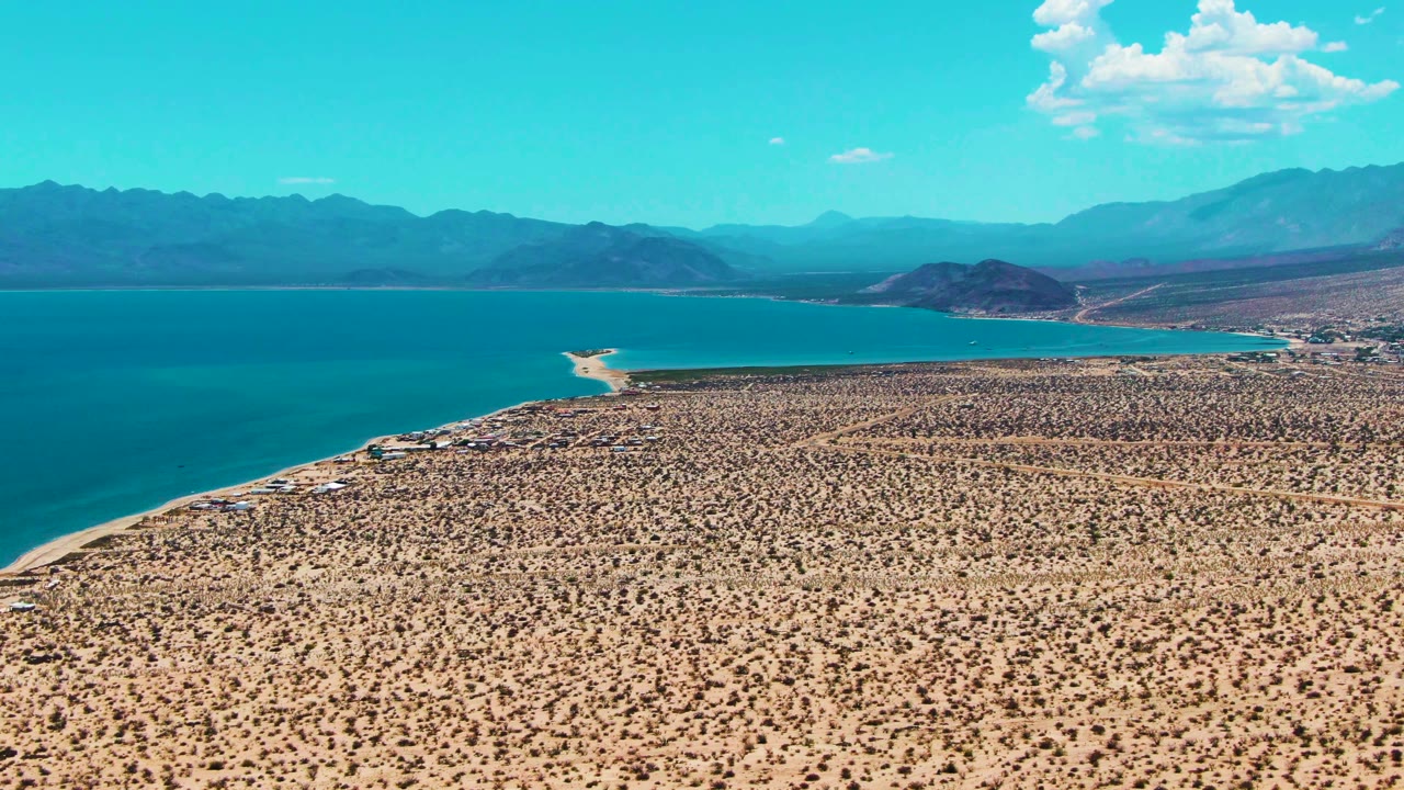 Aerial coastline view in Bahia De Los Angeles with arid terrain in the foreground, water stretching across the left side, mountains beyond, and no visible people.