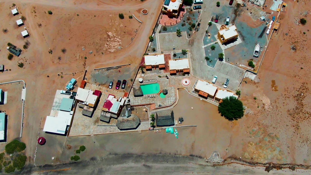 Overhead frame of a Bahia De Los Angeles desert residential area with low buildings, a green pool, sandy lots, parked cars, and no visible people.
