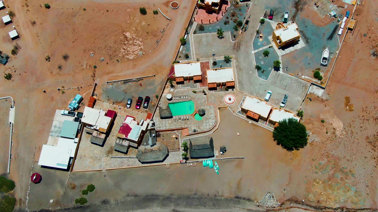 Top-down Bahia De Los Angeles view with pale roofs, red roof sections, parked vehicles, dirt roads, and open desert space with no visible people.