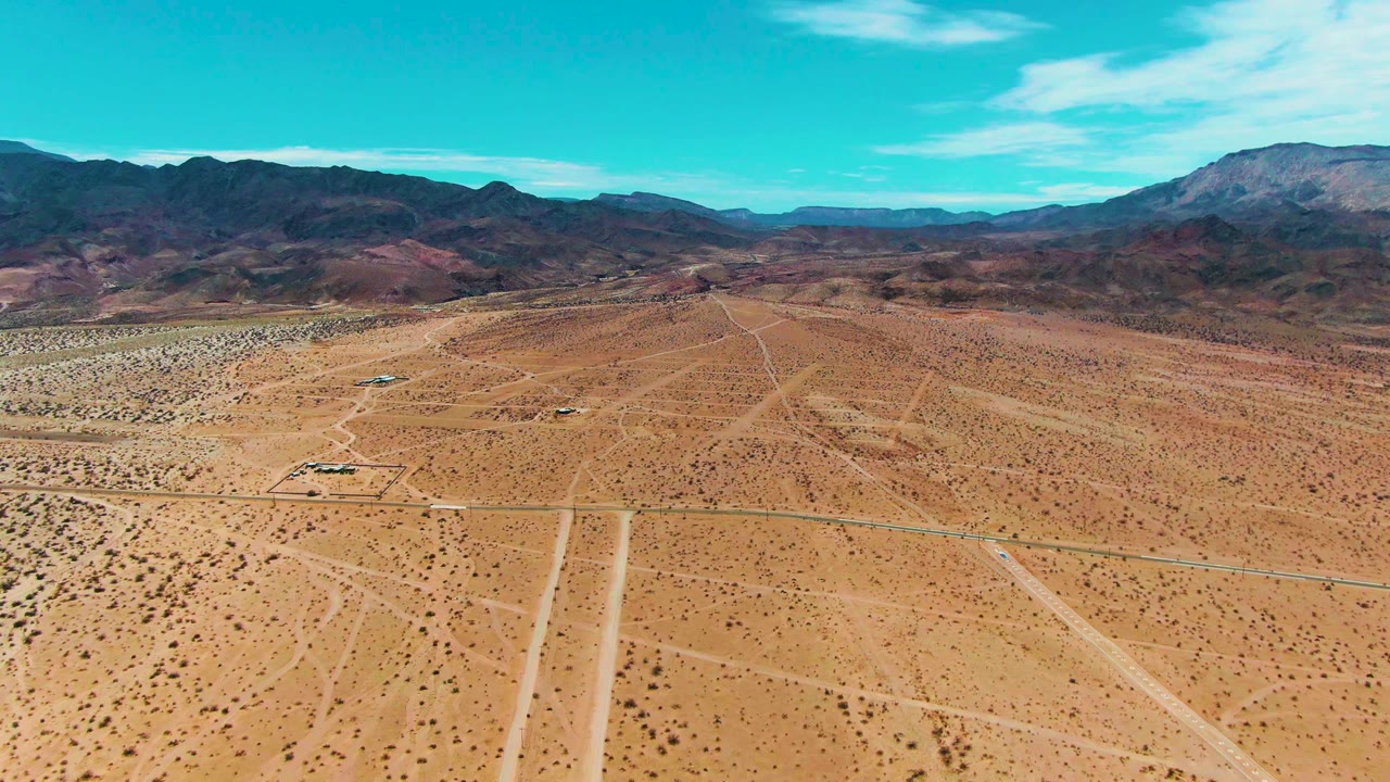 Wide aerial frame in Bahia De Los Angeles showing a small vehicle on the desert road, pale sandy ground, scattered brush, dark mountains, and no visible people.