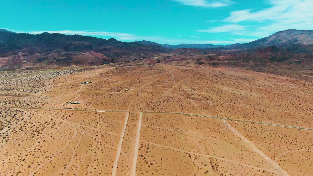 Drone-style view over Bahia De Los Angeles desert with the vehicle farther along the road, low scrub patterns, intersecting dirt tracks, mountains, and no visible people.