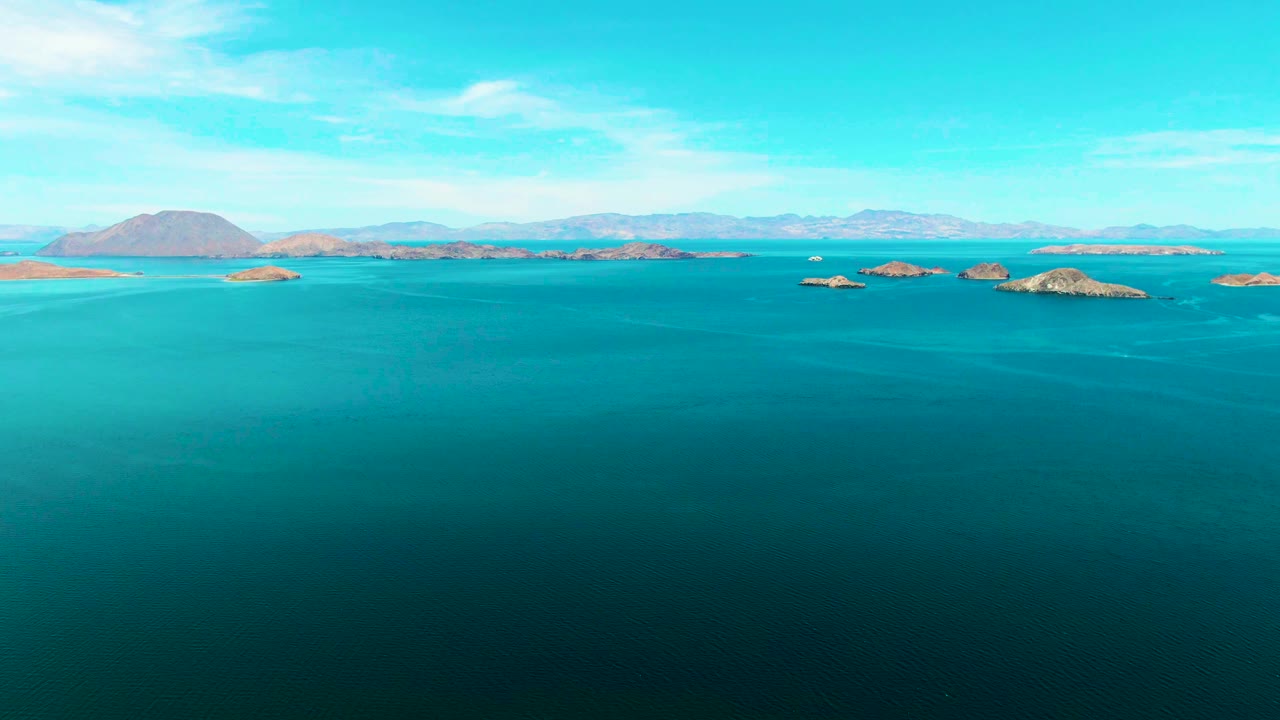 Opening frame of Bahia De Los Angeles with blue-green ocean across the foreground, low rocky islands near the horizon, distant mountains, scattered clouds, and no visible people.