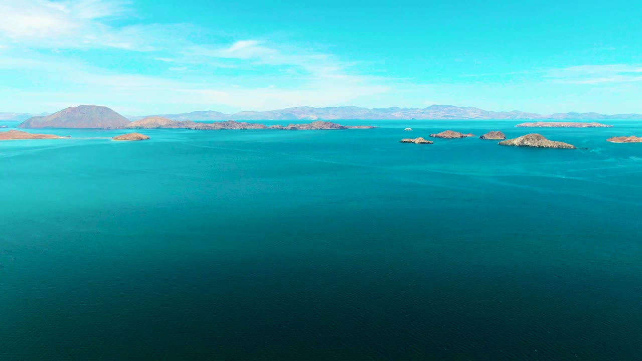 At one second, Bahia De Los Angeles remains framed as open blue water with several rocky islands across the middle distance, pale mountains beyond them, thin clouds, and no visible people.