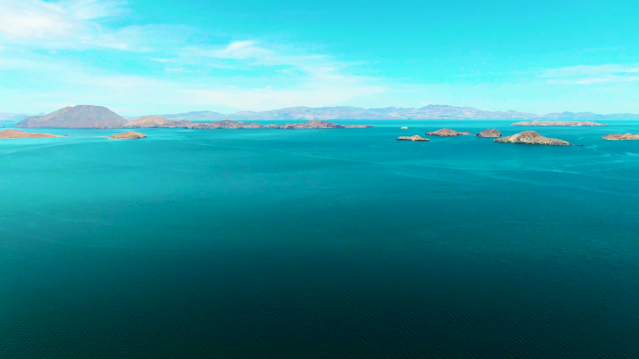 Second frame of the Bahia De Los Angeles view with calm blue sea dominating the lower frame, small rocky islands across the midground, pale mountains beyond, and no visible people.