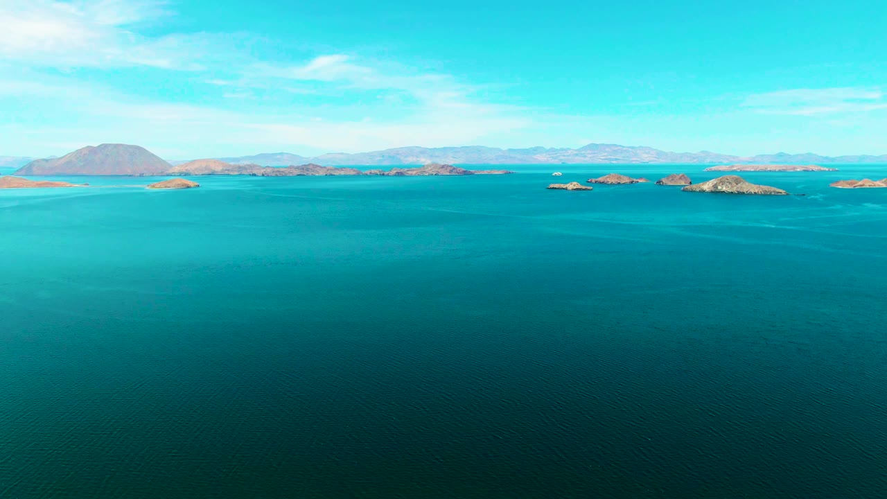 Opening frame of Bahia De Los Angeles, Mexico, showing wide turquoise water, scattered rocky islands on the horizon, distant mountains, thin clouds, and no visible people.