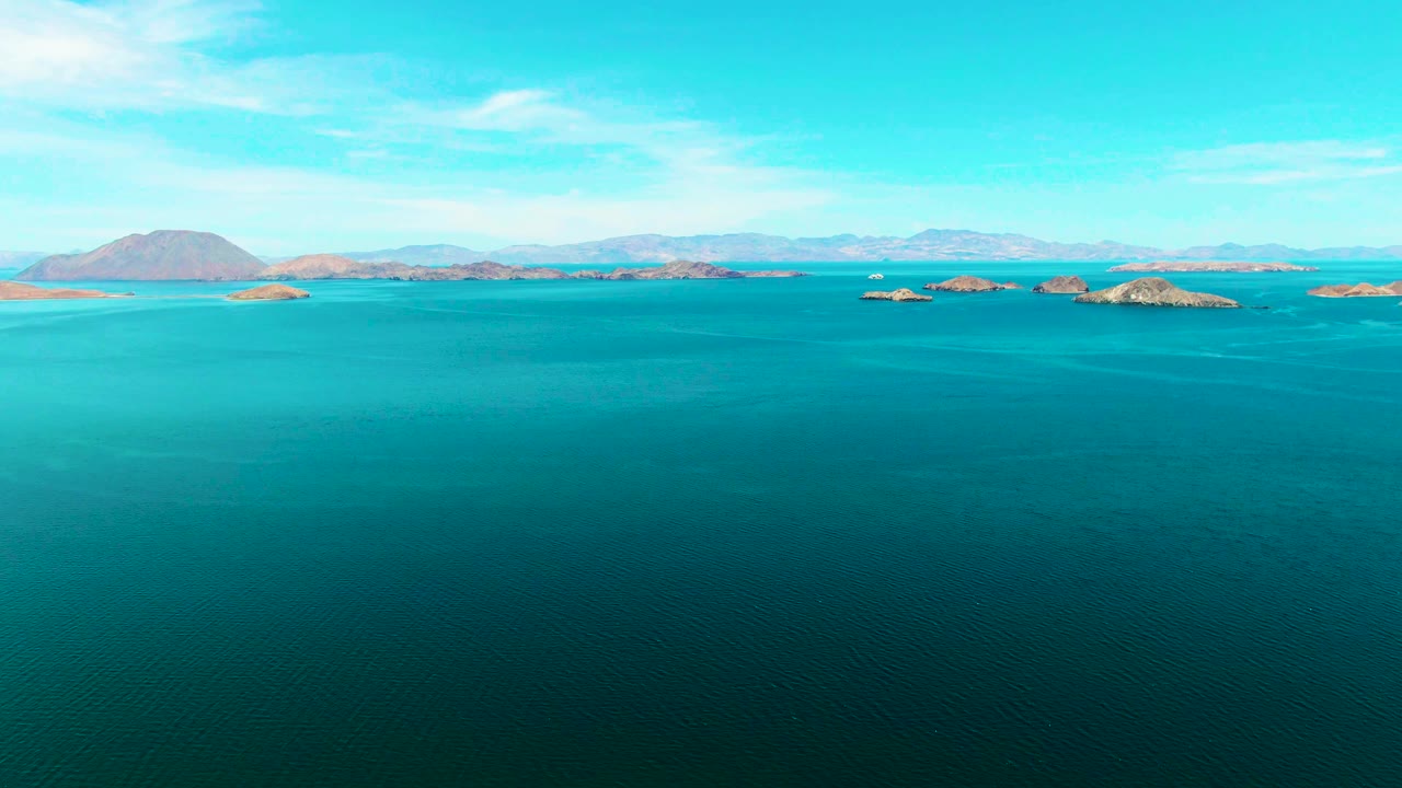 Third frame shows the same Bahia De Los Angeles aerial composition, with broad dark teal water in front, low island clusters across the middle distance, pale mountains beyond, and no visible people.