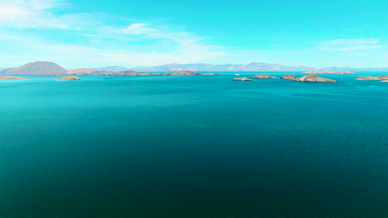 Wide aerial composition over Bahia De Los Angeles shows textured ocean surface, dry island ridges, distant mountains, and no visible people.