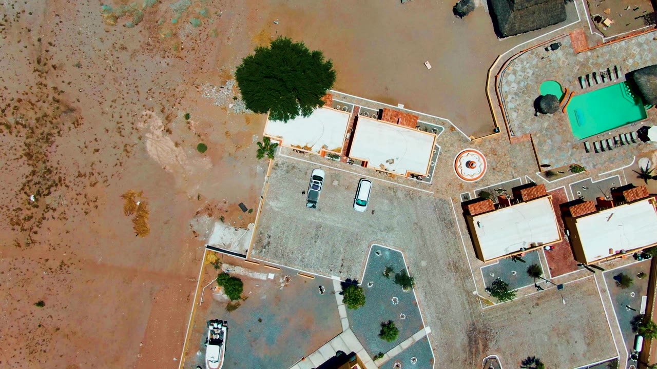Overhead drone frame in Bahia De Los Angeles showing brown floodwater spread around flat-roof buildings, a concrete parking area, parked vehicles, trees, and a turquoise pool, with no visible people.
