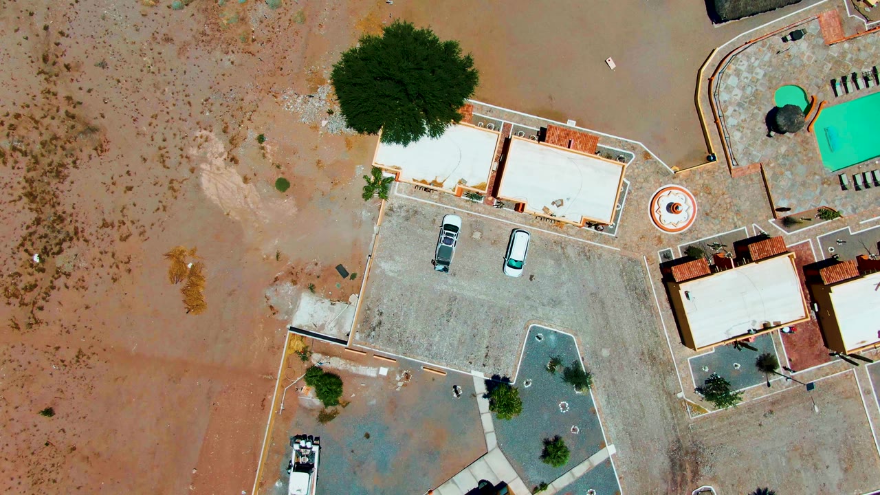 Aerial frame in Bahia De Los Angeles showing muddy floodwater across sandy ground beside homes, parked cars, trees, a circular patio feature, and a bright pool area, with no visible people.
