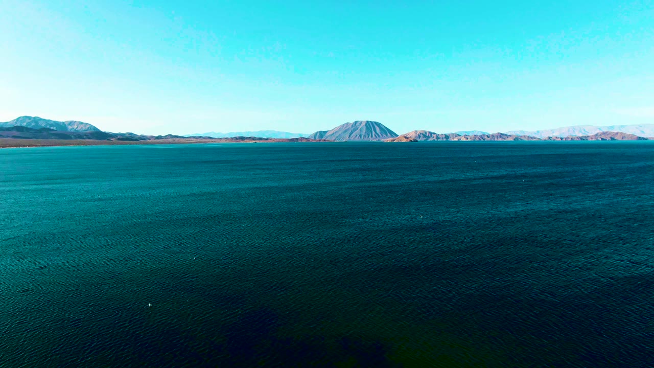 A small white boat is visible on the blue water in Bahia De Los Angeles, with pale mountains across the horizon and no visible people.