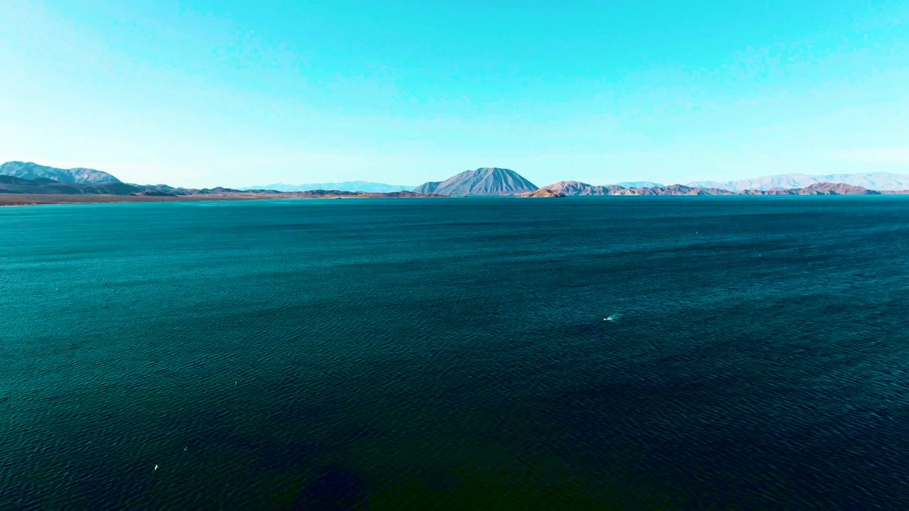 Mid-clip aerial-style view of Bahia De Los Angeles water with a conical mountain near the horizon, pale ridges beyond it, and no visible people.