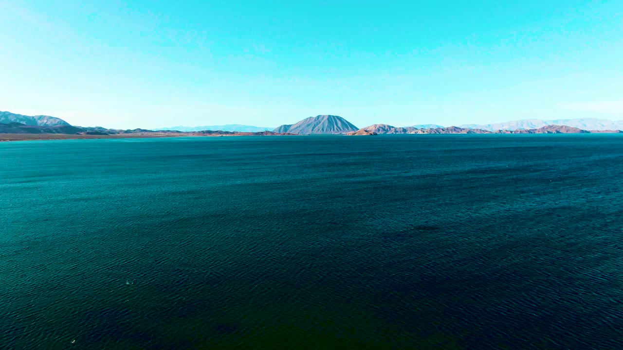 Blue water in Bahia De Los Angeles fills most of the frame, with a low mountain range and clear sky in the background and no visible people.