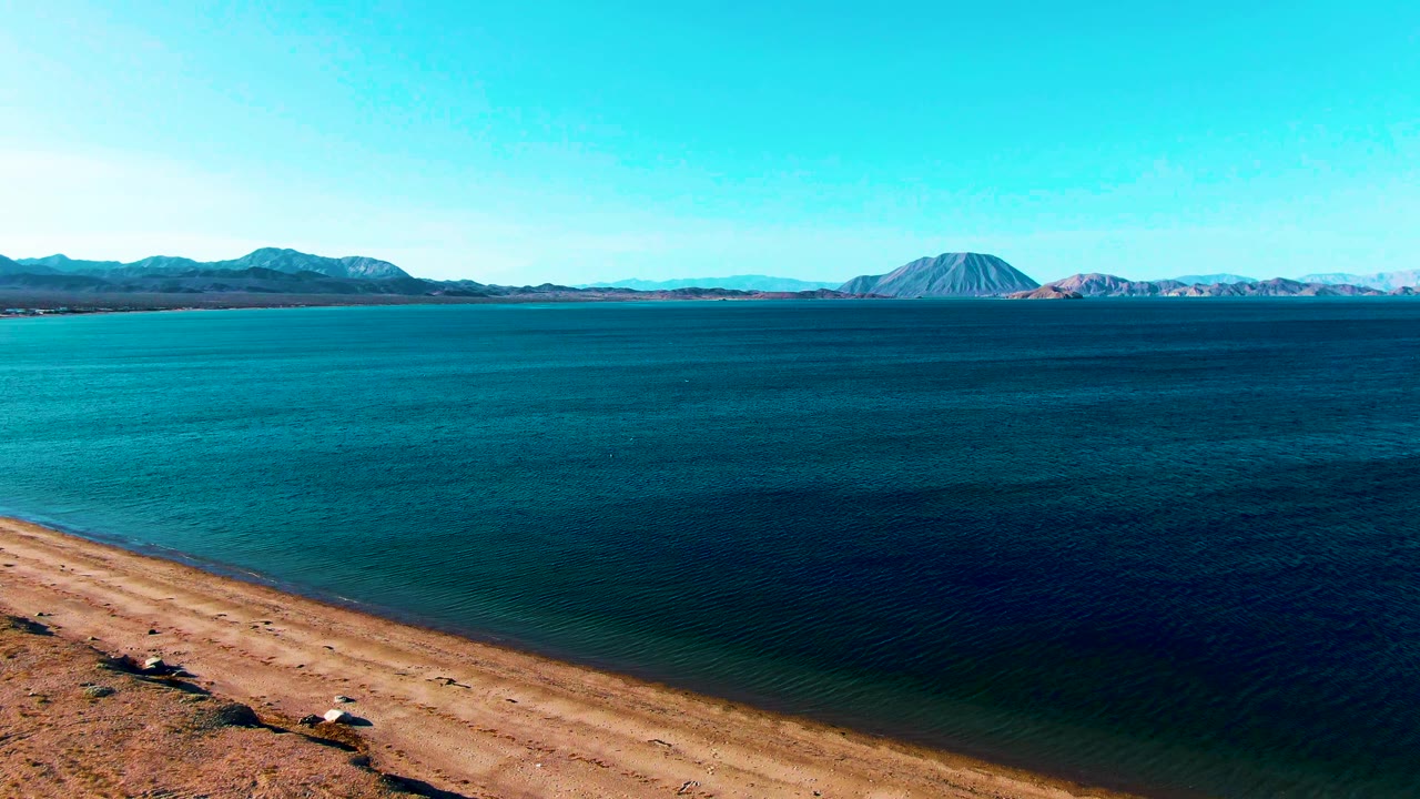 Sandy shore, blue water, and distant mountains under clear sky