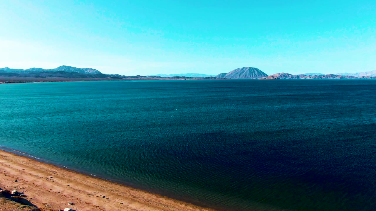 Aerial frame from Bahia De Los Angeles with the sandy edge lower in view, turquoise shallows shifting into darker water, distant mountains, and no visible people.