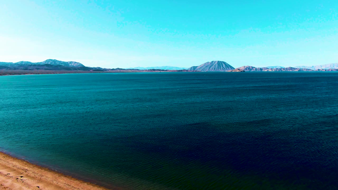 Bahia De Los Angeles aerial view with the shoreline nearly gone from the lower frame, broad blue water across the middle, distant ridge and rounded peak, and no visible people.
