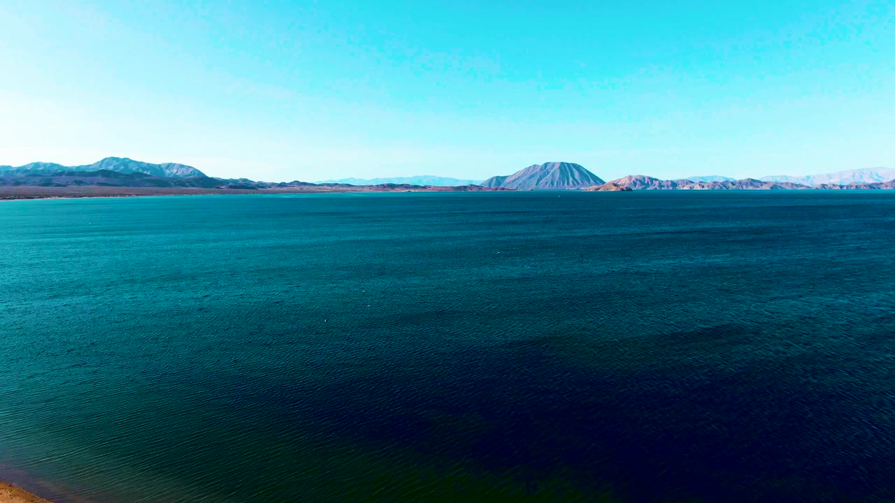 Mid-clip aerial view in Bahia De Los Angeles where blue water fills the foreground and midground, with a low mountain range and rounded peak along the horizon and no visible people.