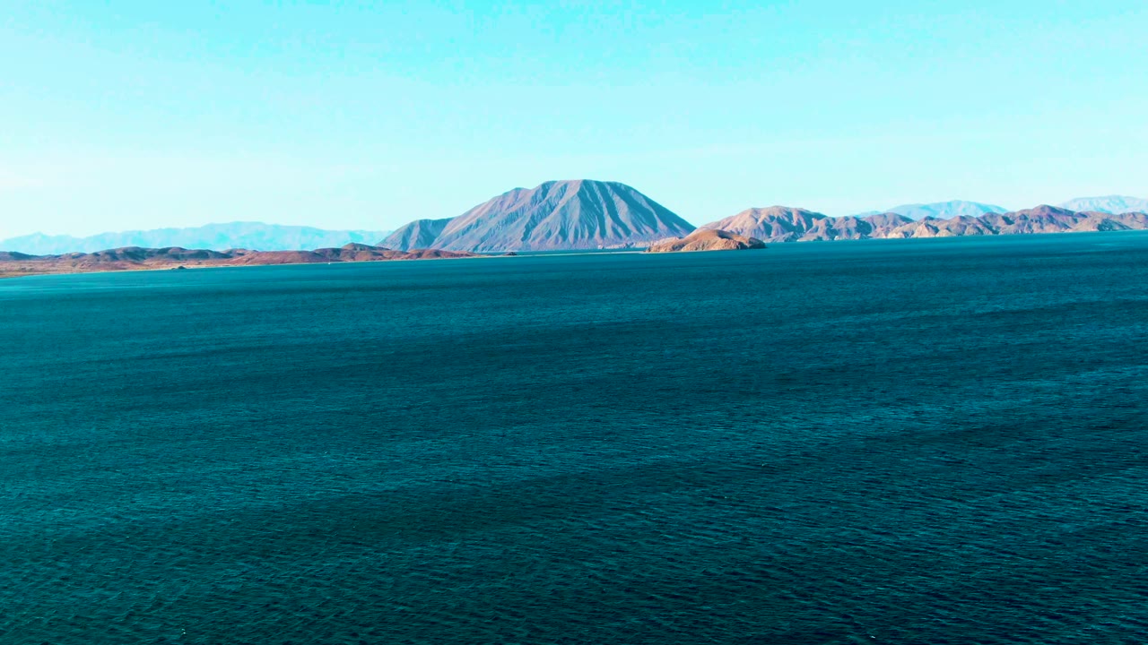 At 0:02, rippled turquoise and deep blue water leads to the brown and gray mountain shoreline of Bahia De Los Angeles, with no visible people.