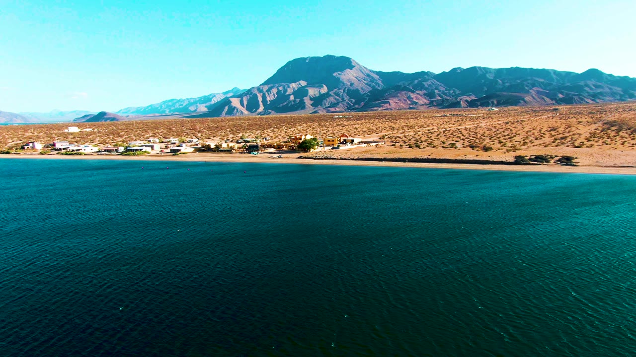 A wide aerial frame of Bahia De Los Angeles shows teal ocean in the foreground, a thin sandy shore with distant buildings, arid mountains, bright sky, and no visible people.