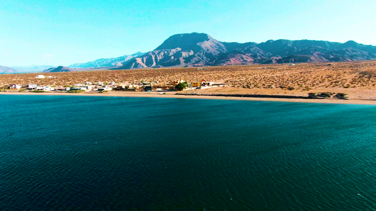 At 0:01, the Bahia De Los Angeles aerial view keeps the blue water dominant, with the sandy beach, low buildings, dry ground, mountains, and no visible people beyond it.