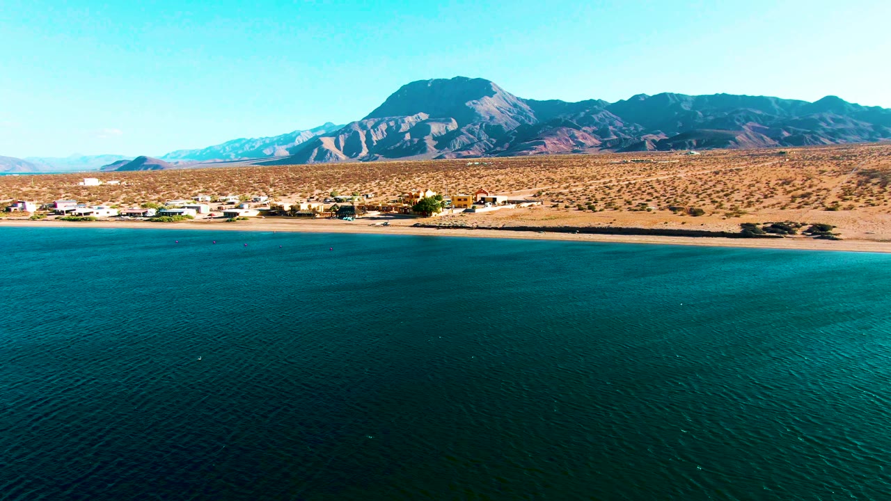 At 0:02, turquoise water stretches toward a narrow Bahia De Los Angeles beach, with scattered settlement buildings, dry brushland, mountains, and no visible people.