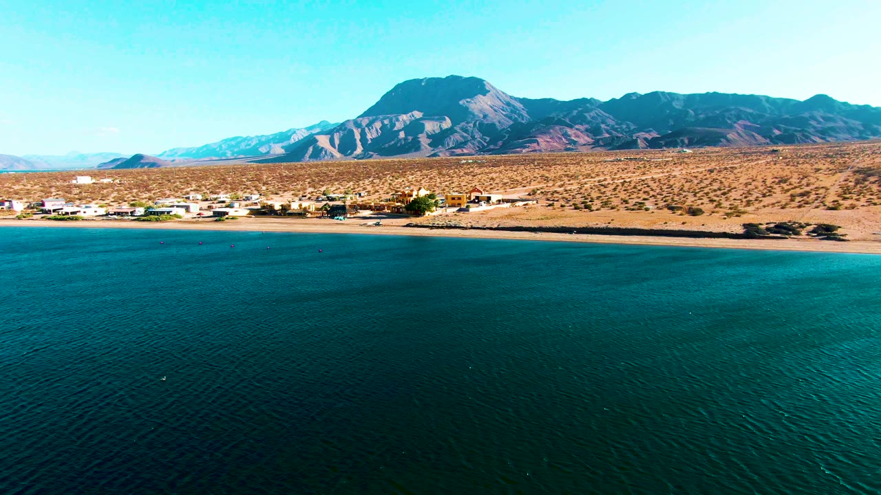 Wide coastal desert aerial with calm ocean and distant mountains.