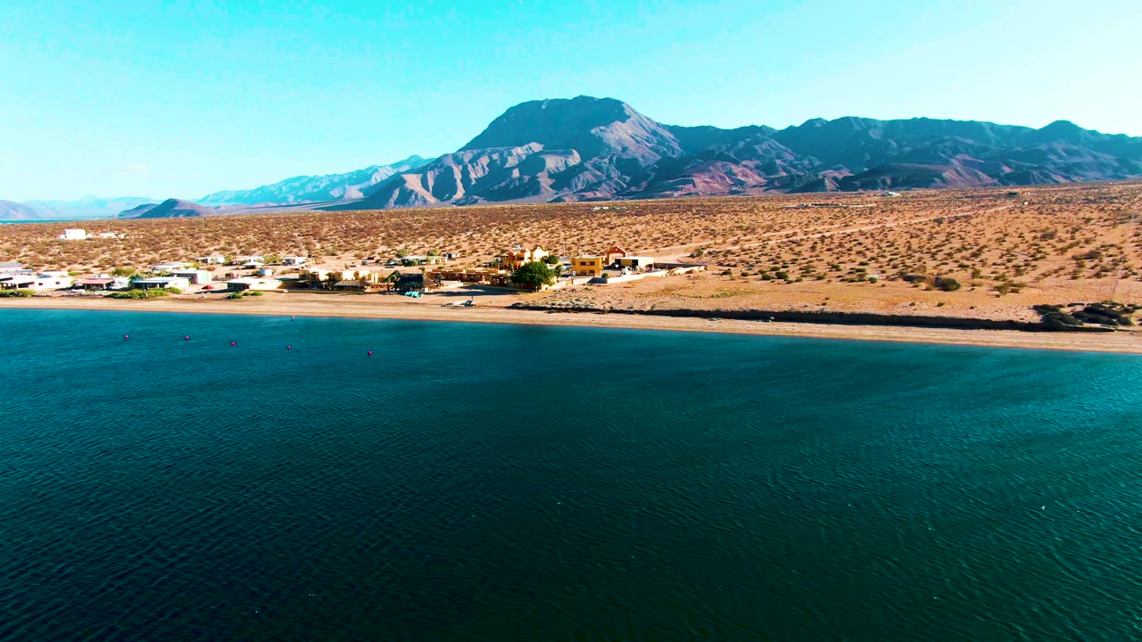 Coastal water, sandy shore, low buildings, and mountain backdrop