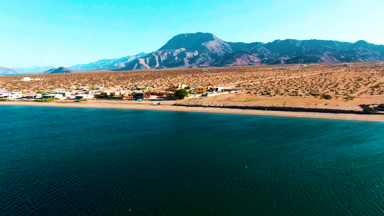 Wide Bahia De Los Angeles aerial with textured teal water across the foreground, pale beach and shoreline buildings beyond it, open desert plain, mountain ridge, and no visible people.