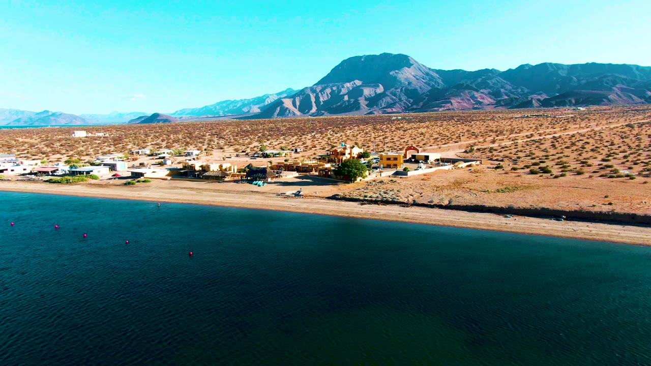 Aerial frame over Bahia De Los Angeles showing calm dark water in front of a sandy beach, a small coastal settlement, dry desert ground, and mountains beyond, with no visible people.