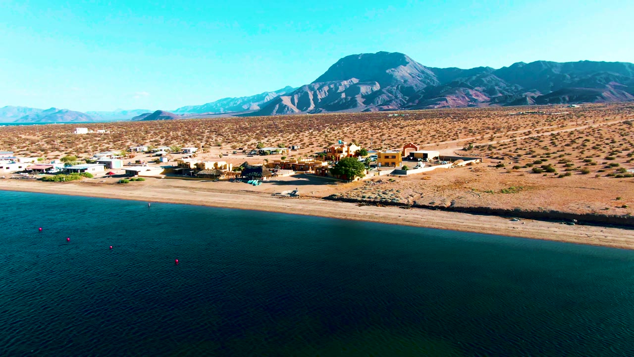 Bahia De Los Angeles appears in a wide aerial composition with calm water across the lower frame, a narrow sandy shore, low buildings near the beach, desert terrain, and mountains under clear sky, with no visible people.