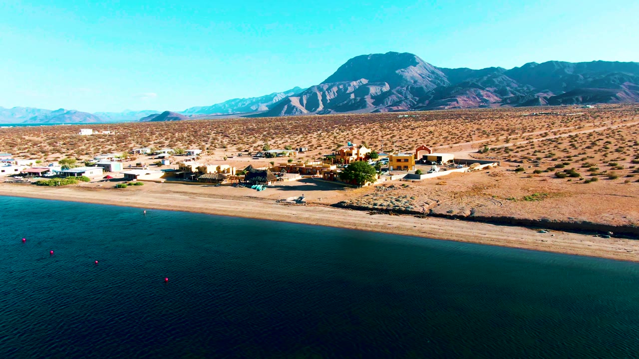 A drone view over Bahia De Los Angeles shows the dark water band, light sand beach, scattered settlement buildings, open desert scrub, and a broad mountain ridge, with no visible people.
